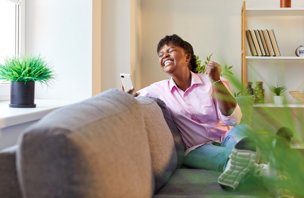 Excited young cheerful happy girl using mobile phone while sitting on sofa at home.