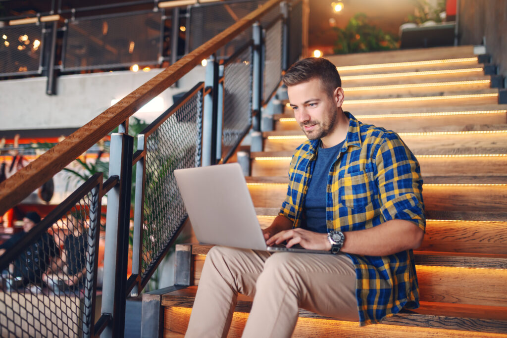 Smiling Caucasian handsome man in plaid shirt sitting on stairs in cafe and using laptop for work.