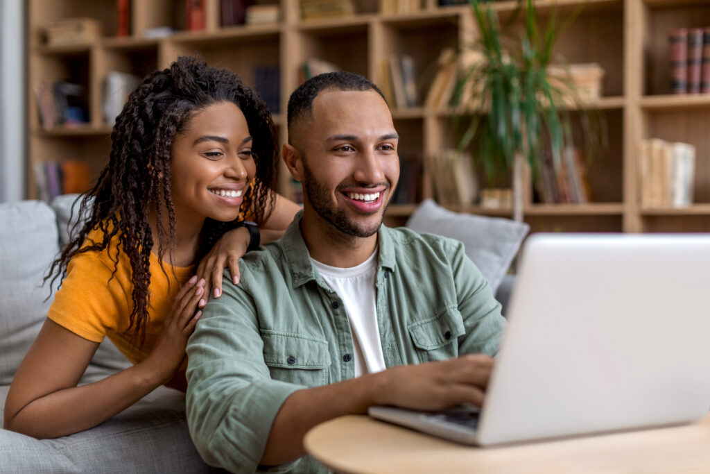 Happy african american spouses sitting by couch with laptop, looking for new furniture online, websurfing, copy space