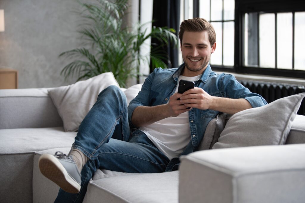 Attractive smiling young man wearing casual clothes sitting on a couch at the living room, using mobile phone