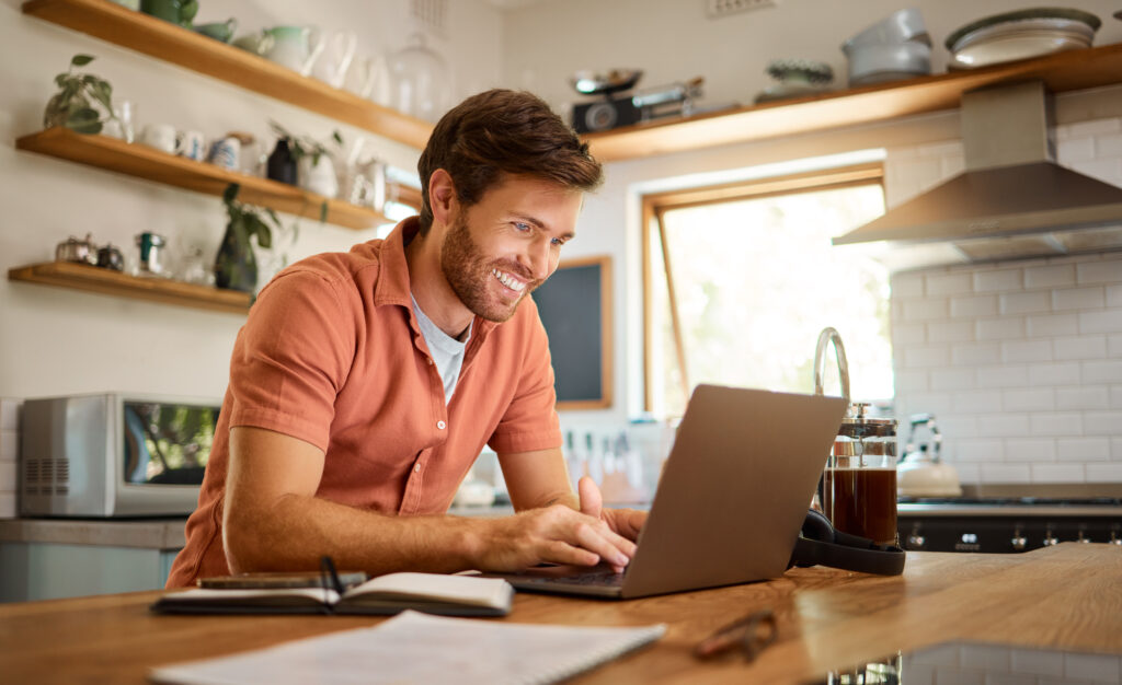 Smile, laptop and remote work, happy man at kitchen counter with app for email, social media or networking for online job. Technology, communication and freelancer on internet writing blog or article