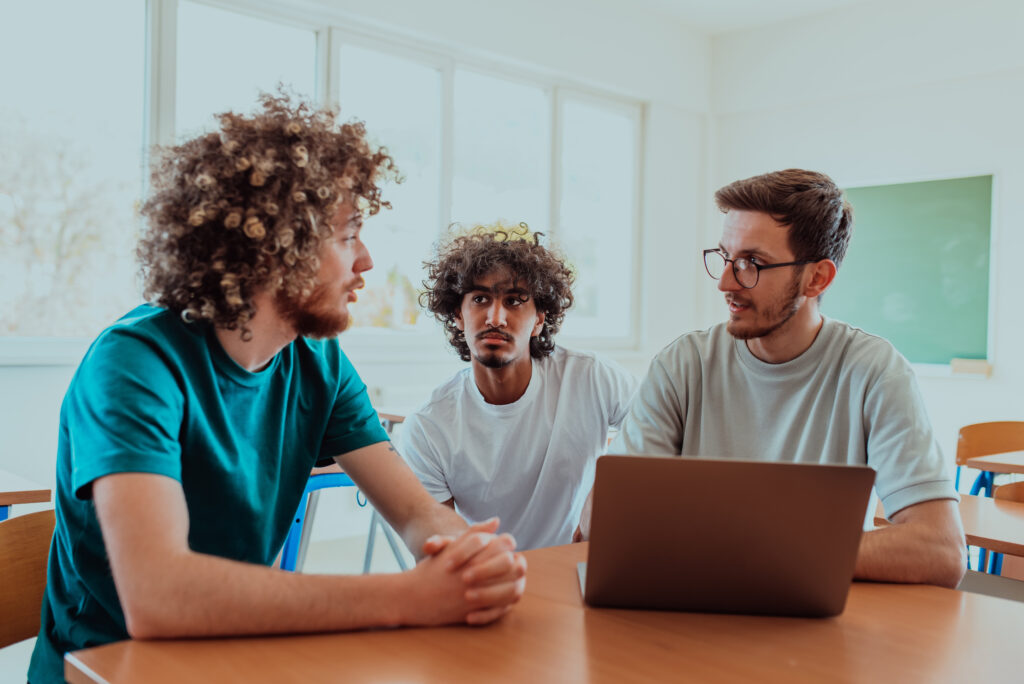 Group students including one African-American, sit together in a classroom, engaging in a discussion while reviewing information on a laptop