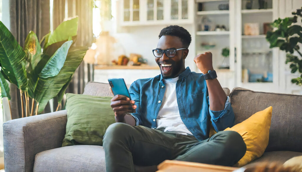 Excited looking young man holding smartphone device. Man making yes gesture with clenched fist. Man with happy and satisfied expression looking at smartphone screen.