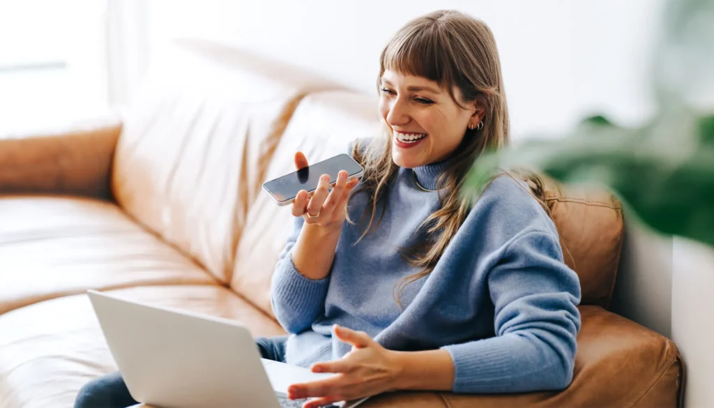 Jeune femme souriante en train de faire une dictée vocale avec son téléphone, assise sur un canapé, tout en travaillant sur un ordinateur portable